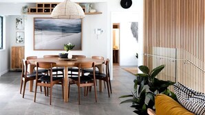 A dining area with a wooden table and chairs, a large painting on the wall, and a staircase with wooden railings. - 5 BDR - 250m to Beach - Outdoor Spa - Book 3 Nights, Pay for 2 until 24 September 2026 (Hyams Beach)