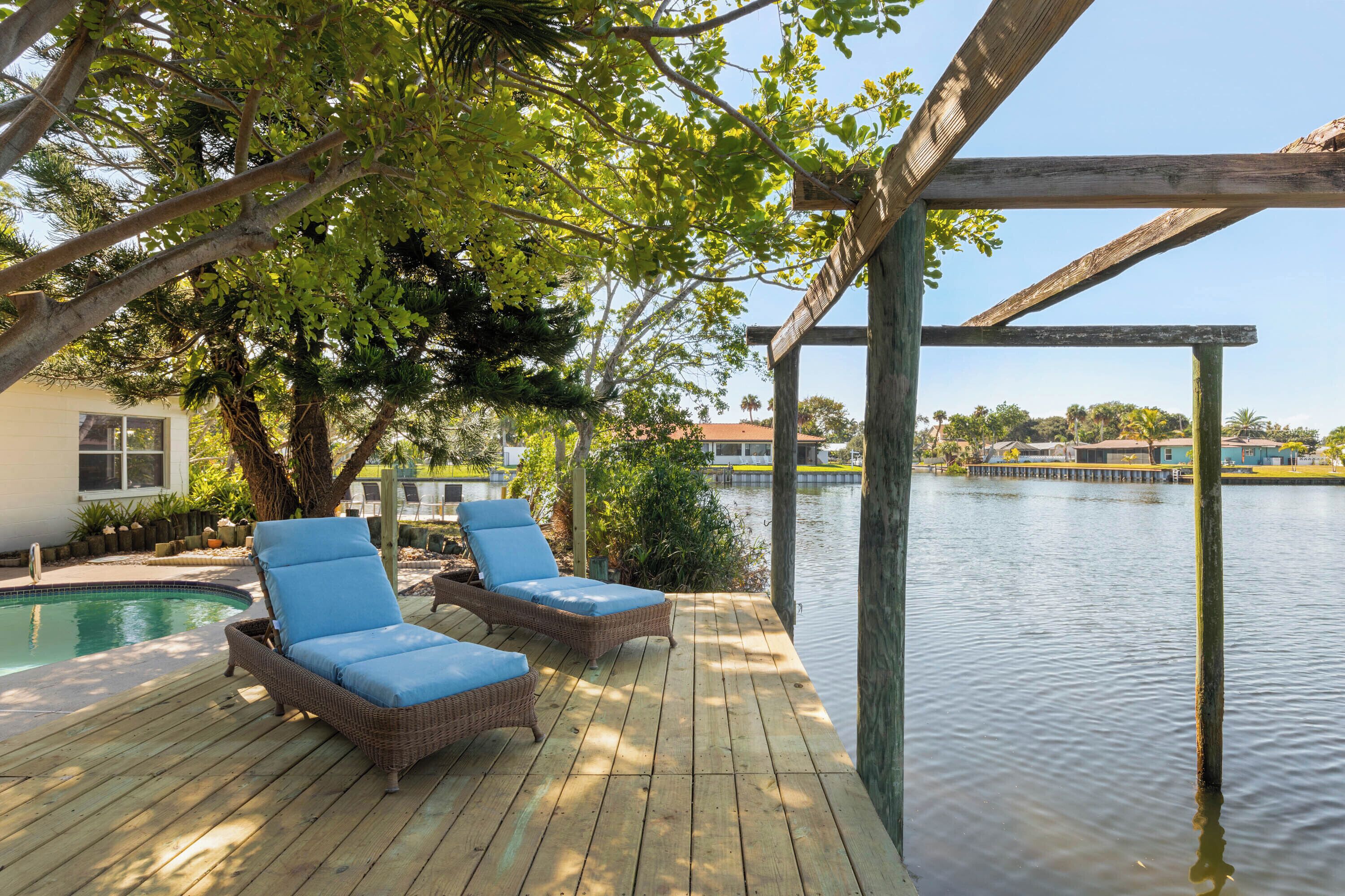 Terraza de madera con dos sillones de mimbre, piscina y vista a un cuerpo de agua.