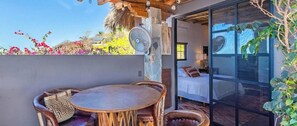 Eine Terrasse im Freien mit einem runden Holztisch und Stühlen, einem Ventilator und Blick auf ein Schlafzimmer mit Bett und Fenster.