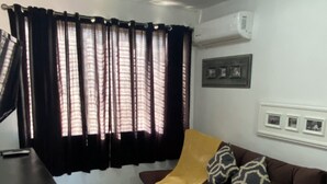 Una sala de estar con un sofá marrón, una manta amarilla y cojines estampados. Hay una mesa de centro negra, una alfombra gris y una ventana con cortinas negras.