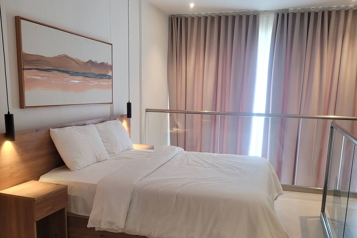 A modern bedroom with a wooden headboard, a large bed with white linens, a nightstand, a bedside table, a wall-mounted lamp, a painting, and a view of the outside through a window with curtains.