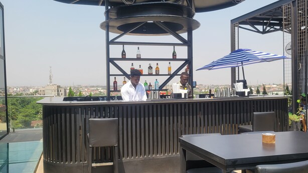 A bar area with a bartender, a counter with various bottles, and outdoor seating. - RT properties at Belrose Place (Accra)