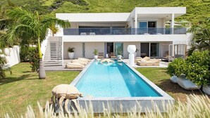 Uma casa moderna de dois andares com piscina, rodeada por vegetação exuberante e colinas.