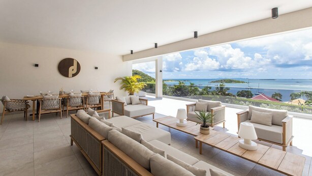 Ampla sala de estar com uma grande janela com vista para o oceano, mobiliada com sofás, uma mesa de jantar e uma mesa de centro.