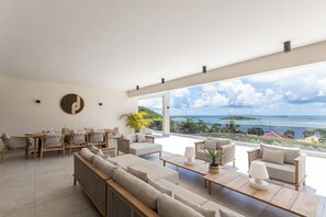A spacious living area with a large window overlooking a scenic ocean view, furnished with sofas, a dining table, and a coffee table.