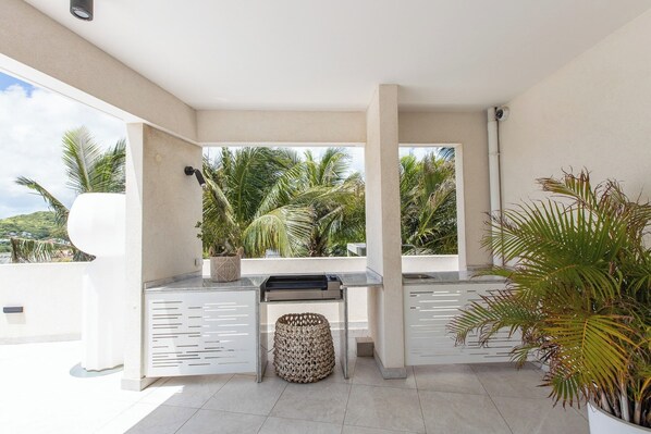 A modern outdoor kitchen area with a built-in grill, a seating bench, and a large potted plant.