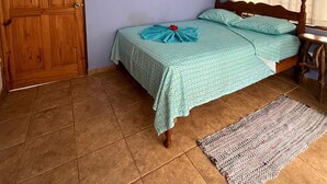 Um quarto com porta de madeira, cama com colcha verde, tapete pequeno e janela com cortinas brancas.