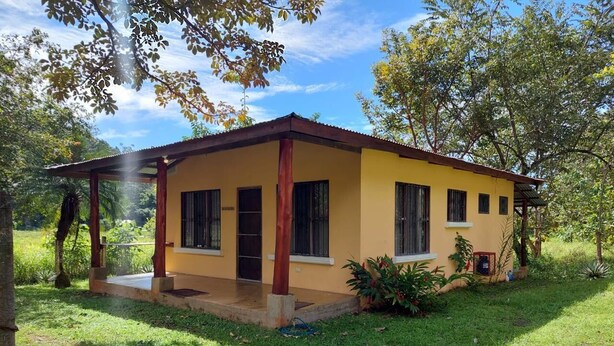 Uma casa amarela térrea com varanda, cercada por vegetação.
