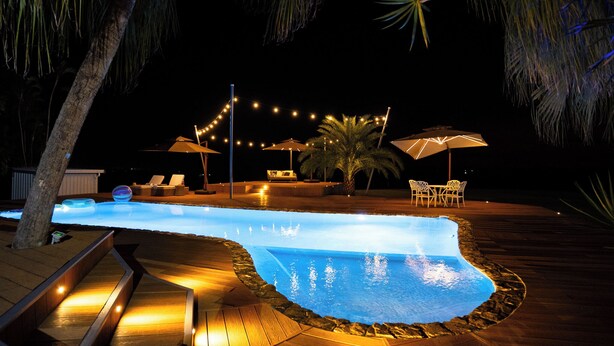 Un área de piscina exterior de noche con muelle de madera, sillas de descanso e iluminación con luces colgantes.