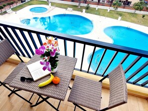 A balcony with wicker chairs, a table with a book, a flower vase, a banana, and an orange. In the background, there are two swimming pools.