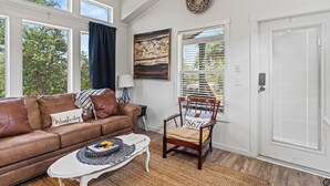 A living room with a brown leather sofa, a wooden chair, a white door, and a large window with blinds. - Hidden Fox Escape - Grey Fox (Hays County)