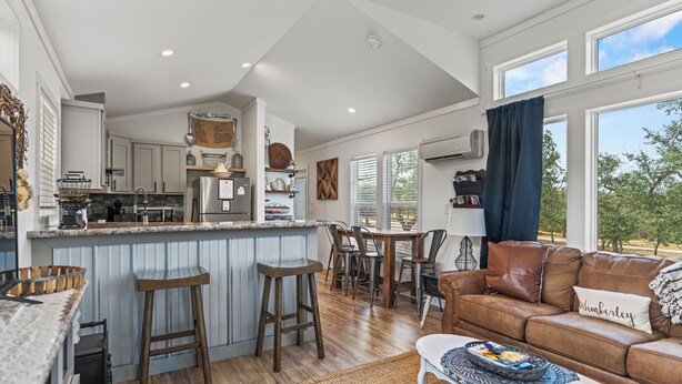 A spacious living area with a kitchen, bar stools, a leather sofa, and a large window offering a view of the outdoors. - Hidden Fox Escape - Grey Fox (Hays County)