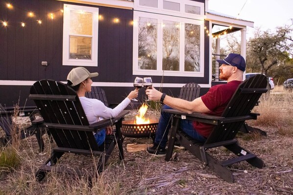 Twee personen die met wijn glazen proosten bij een kampvuur, zittend op Adirondack stoelen, met een huis en lichtjes op de achtergrond.