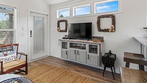 A living room with a TV, grey cabinets, a bench, and a door. - Hidden Fox Escape - Grey Fox (Hays County)