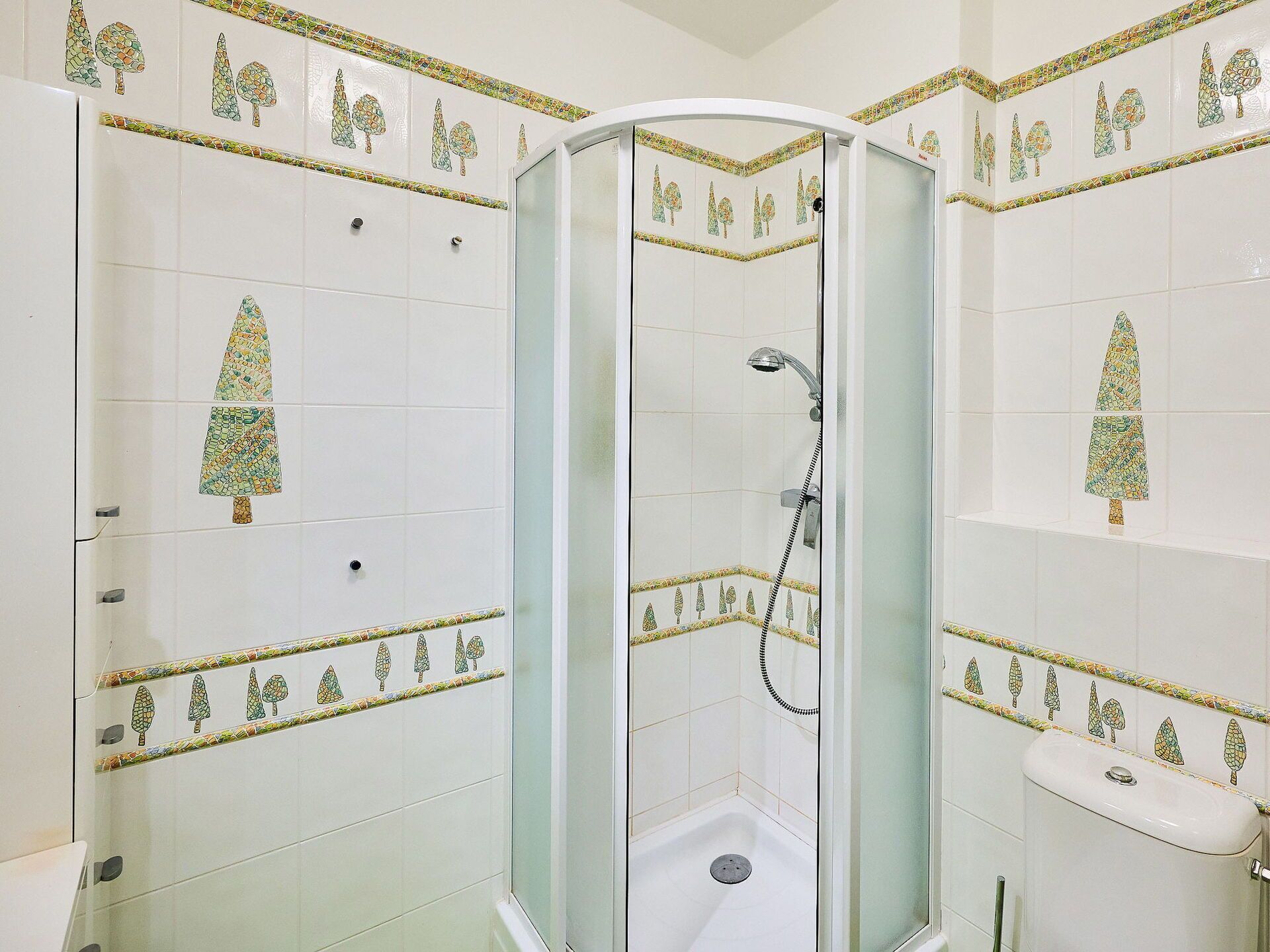 A bathroom with a glass-enclosed shower, white tiled walls, and decorative tree patterns.