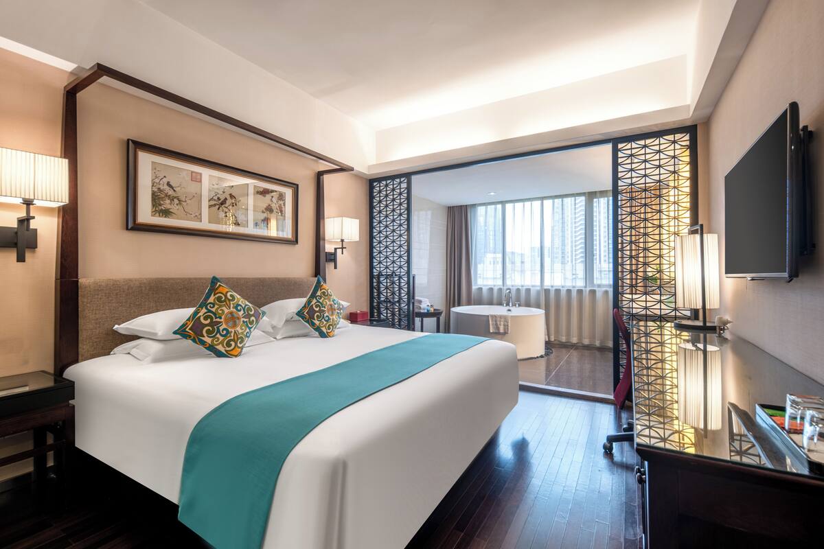 Ein Hotelzimmer mit einem großen Bett, einem Schreibtisch, einem Fernseher und einem Badezimmer mit Badewanne, mit Blick auf die Stadt.