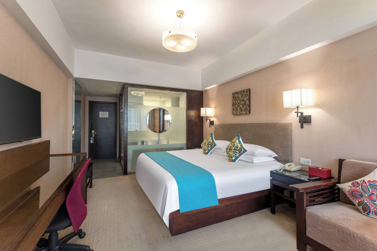Ein Hotelzimmer mit einem großen Bett, einem Schreibtisch mit Telefon, einem Sessel, einem Fernseher und einer Tür zu einem Flur.