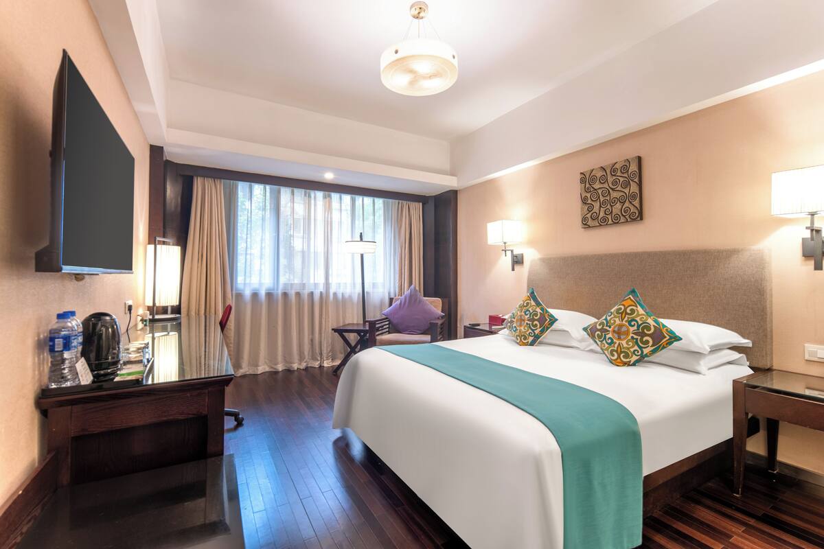Ein Hotelzimmer mit einem großen Bett, einem Schreibtisch mit Flachbildfernseher, einem Sessel, einer Lampe und einem Fenster mit Vorhängen.