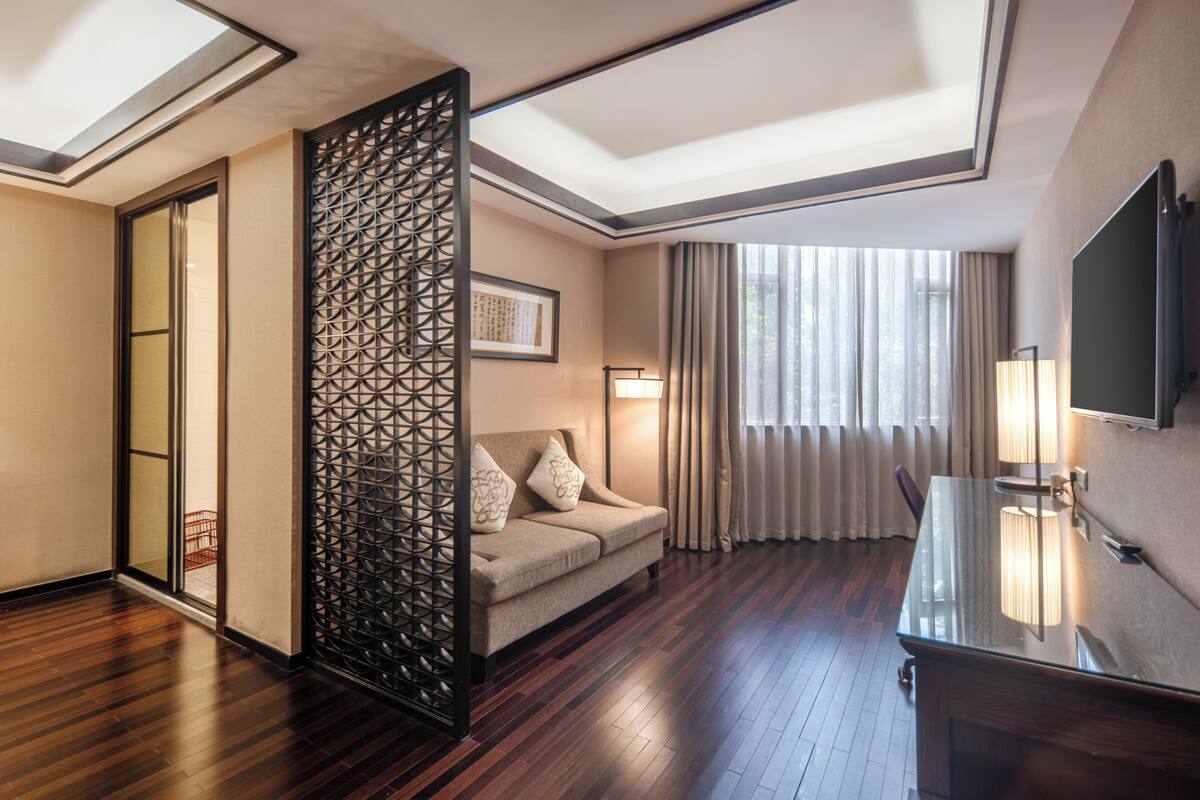 Ein Hotelzimmer mit Holzboden, einer Couch mit Kissen, einem Fernseher, einem Fenster mit Vorhängen und einer Zier trennwand.