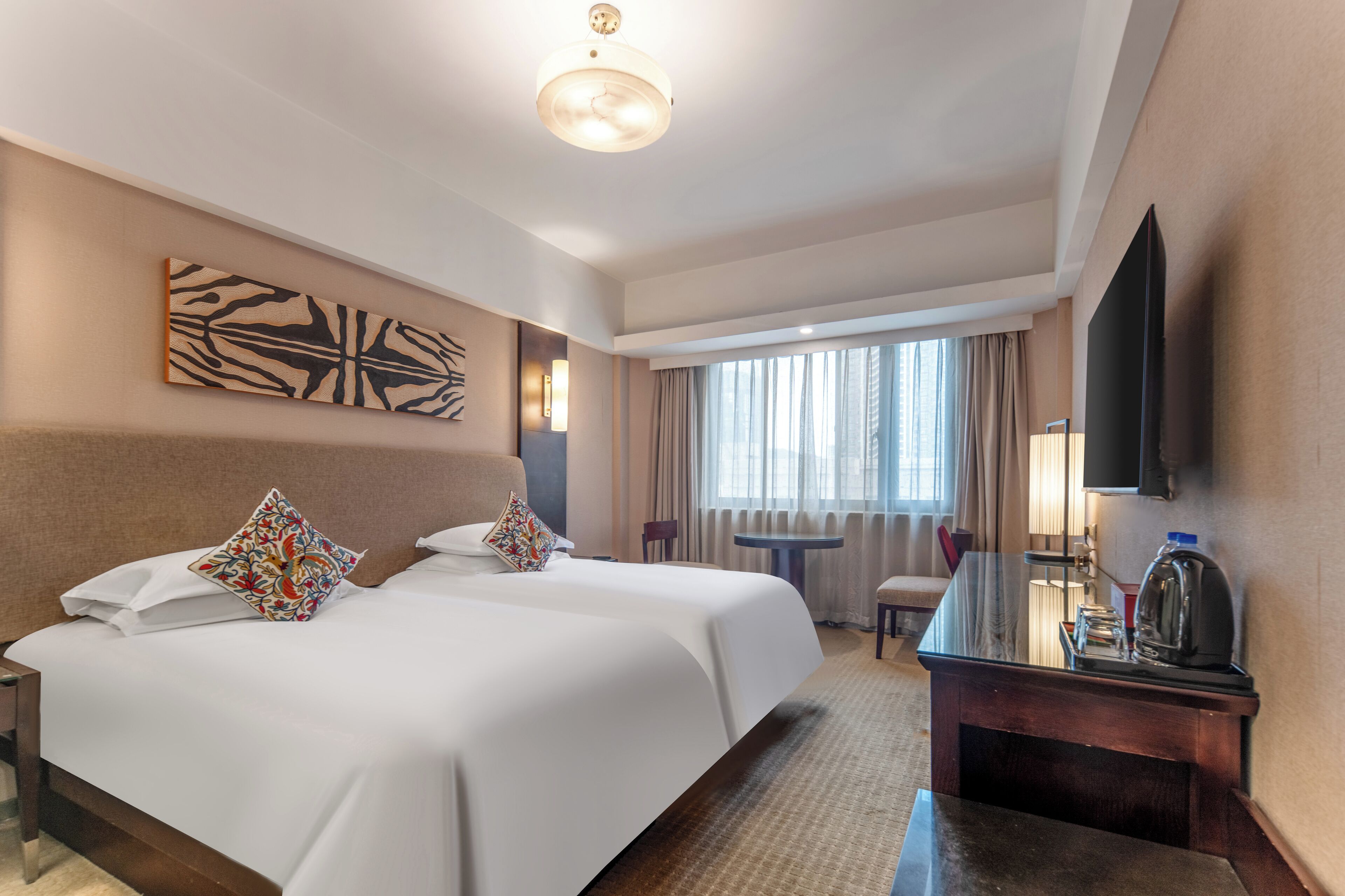 A hotel room with two beds, a desk, a flat-screen TV, and a large window with curtains.