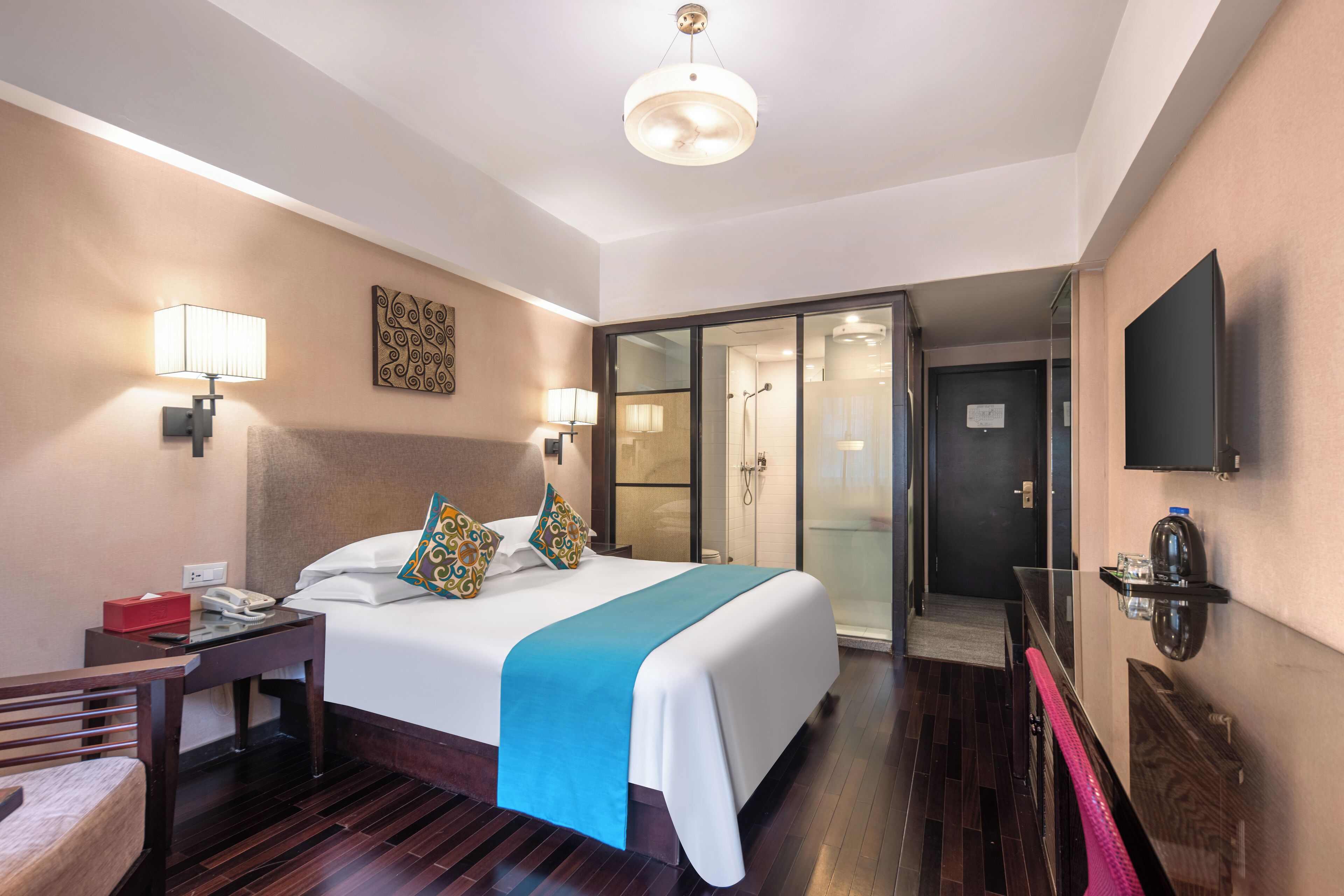Ein Hotelzimmer mit einem großen Bett, Nachttischlampen, einem Flachbildfernseher, einem Schreibtisch, einem Sessel, einem Duschbereich und einer Glastür zum Badezimmer.