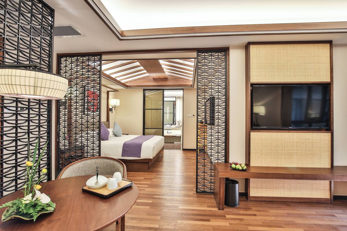 Une chambre d’hôtel avec un plancher en bois, un lit recouvert d’une couverture violette, un téléviseur fixé au mur et une table à manger agrémentée d’un vase de fleurs.