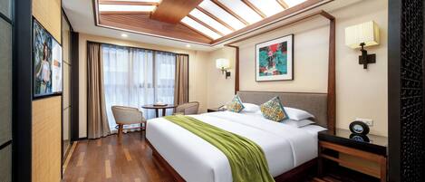 A hotel room with a large bed, a desk, a chair, a window with curtains, a painting on the wall, and a ceiling with wooden beams.