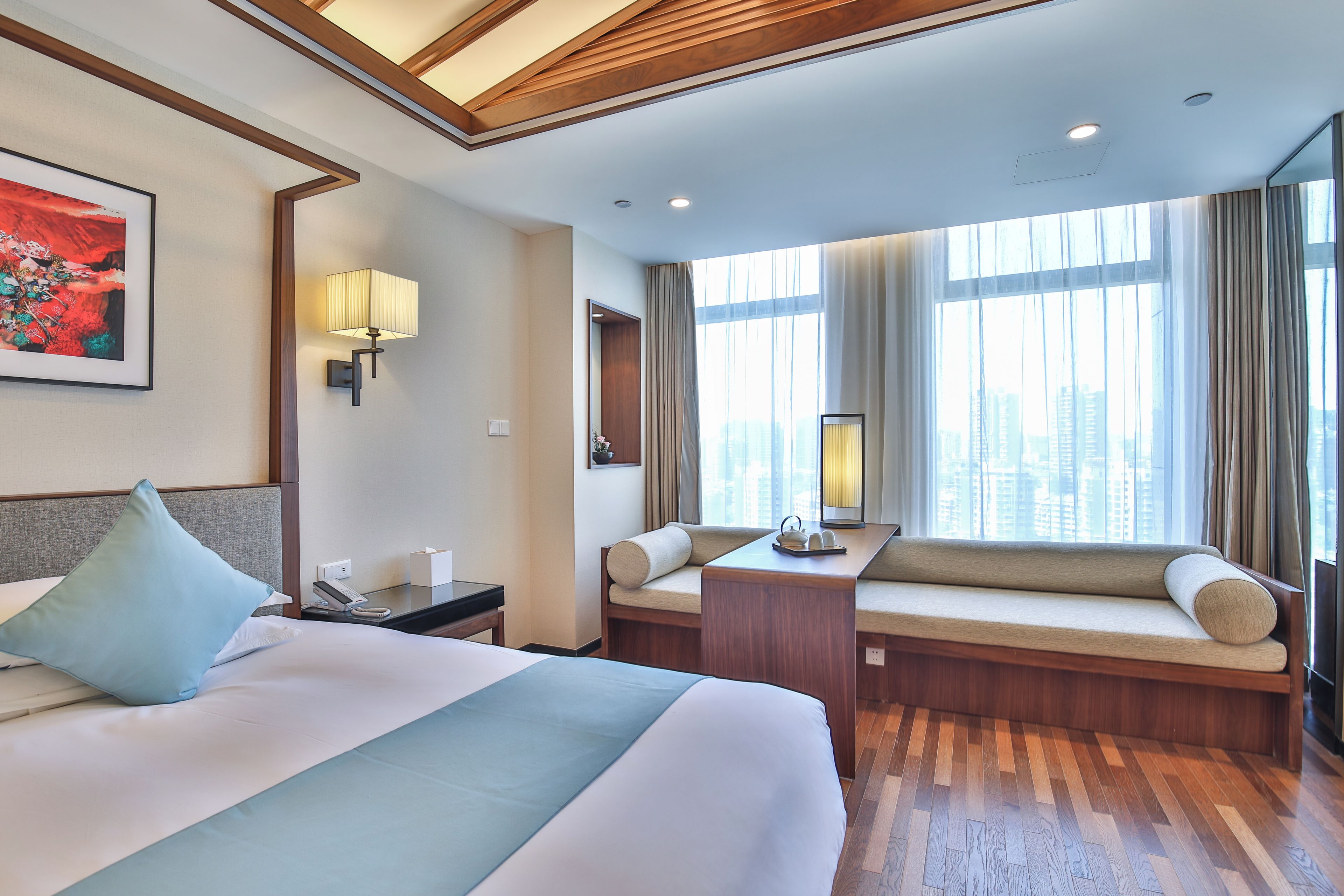 A hotel room with a large bed, a desk, two seating areas with cushions, a painting on the wall, and a city view through the window.