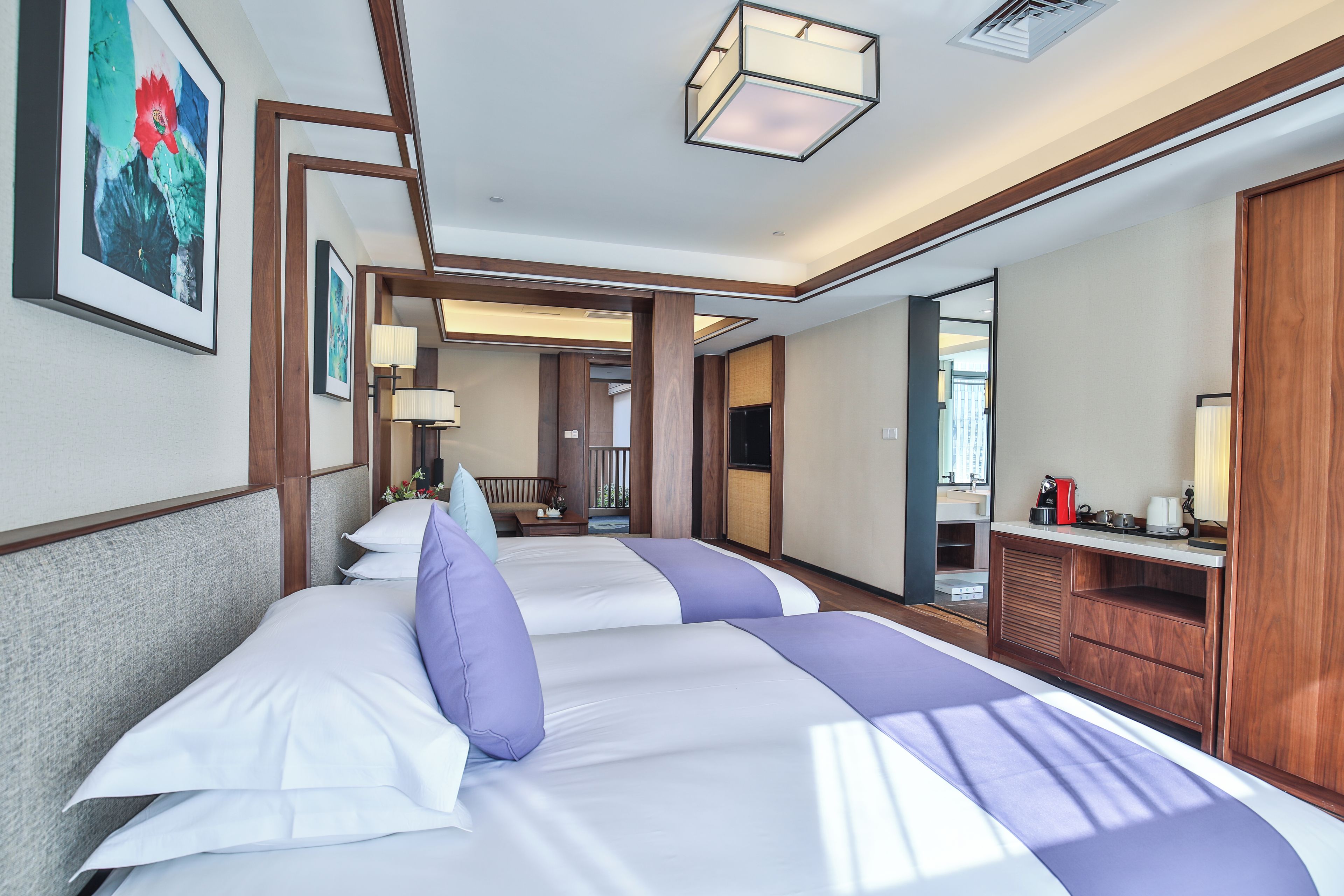 A hotel room with two beds, a wooden wardrobe, a desk with a lamp, and artwork on the wall.