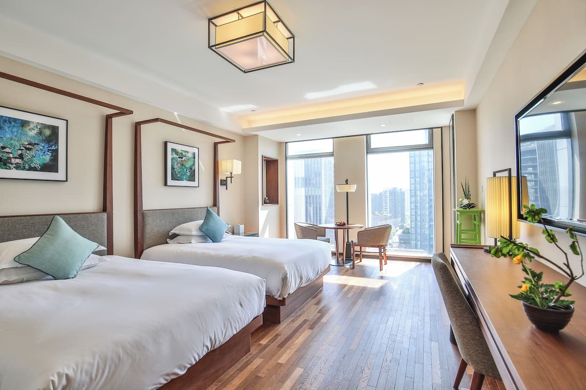 Une chambre d’hôtel avec deux lits, un coin repas et de grandes fenêtres donnant sur la ville.