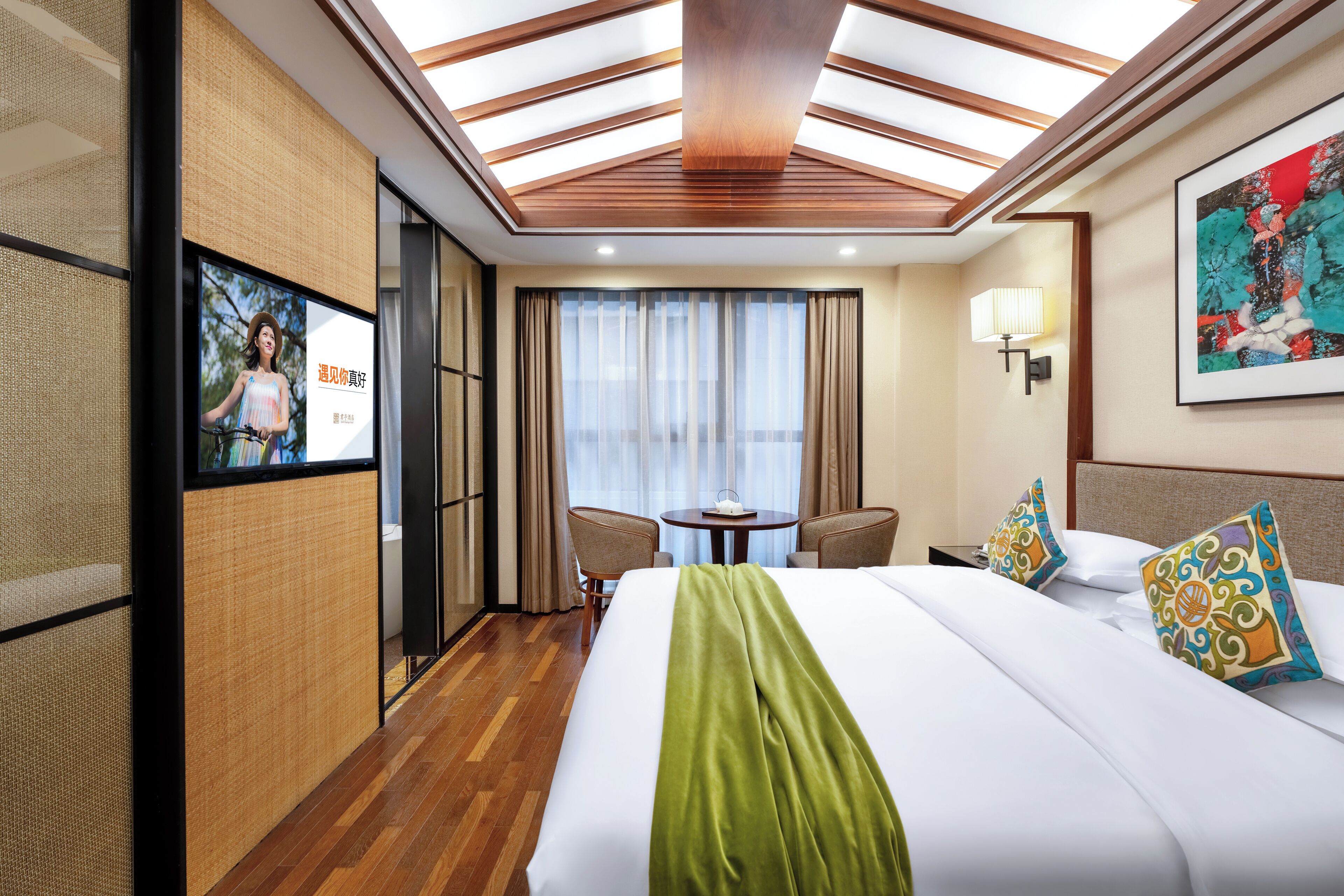 Una camera d'hotel con un letto grande, una scrivania, una sedia, una finestra con tende e un quadro appeso al muro, oltre a un soffitto con lucernari.