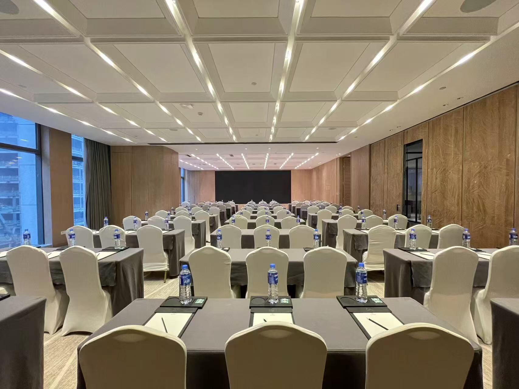 A conference room with rows of tables and chairs, equipped with water bottles, and large windows offering a view of the cityscape.