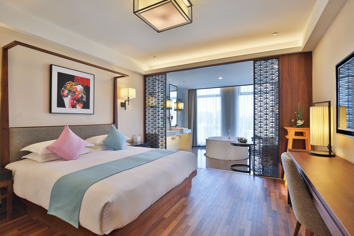 A hotel room with a large bed, a desk, a chair, a bathroom with a bathtub, and a view of the city.