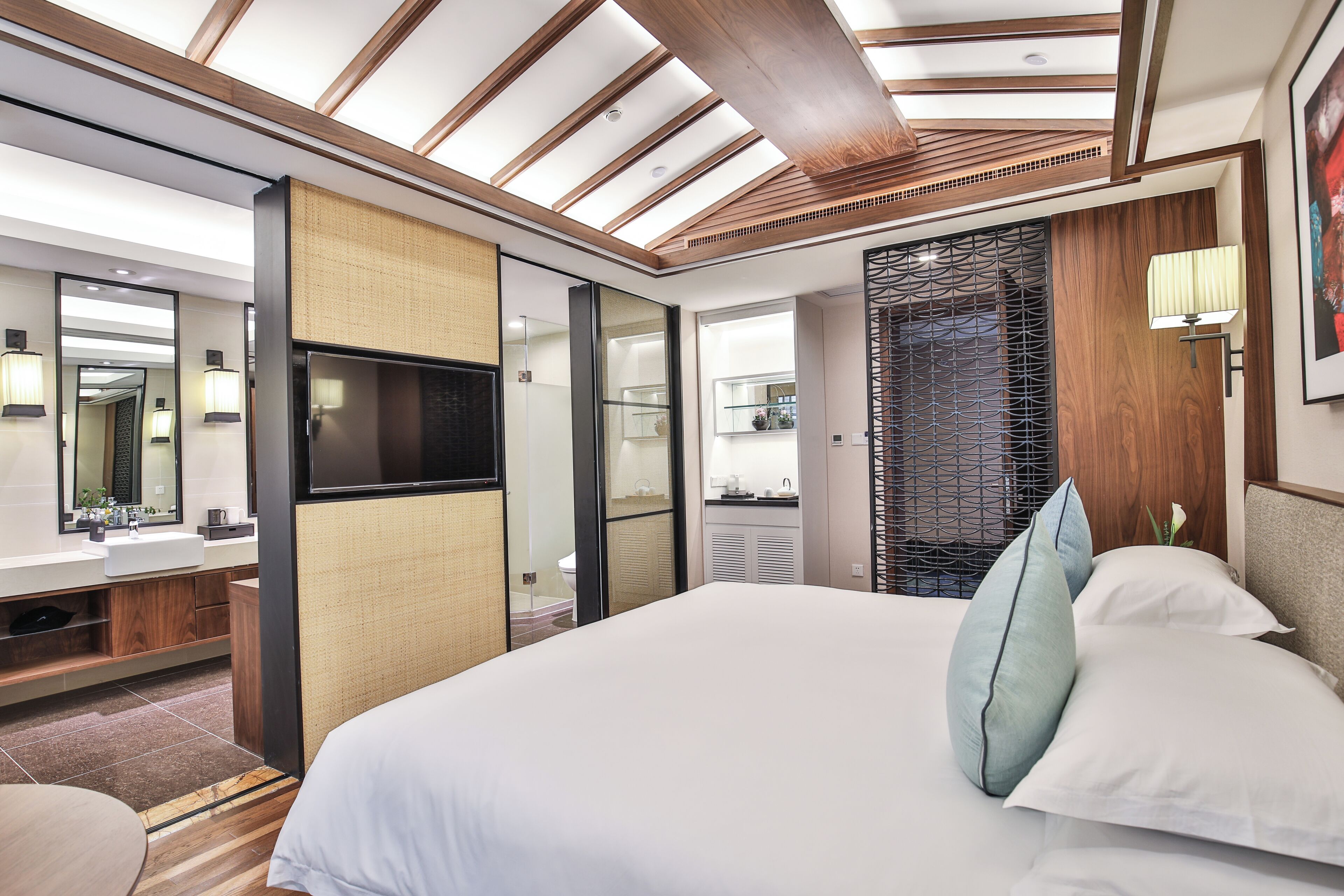 Una habitación de hotel moderna con una cama grande, un televisor de pantalla plana, un baño con espejo y lavamanos, y techo de madera.