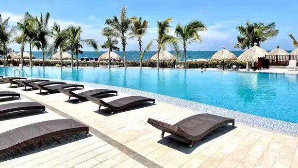 Un espace aménagé au bord de la piscine, avec des chaises longues et des palmiers, donnant sur une piscine à l’eau d’un bleu limpide.