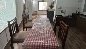Un coin repas avec une table recouverte d’une nappe à carreaux, des chaises en bois, un espace cuisine comprenant un évier et une cuisinière, et une fenêtre avec des stores.
