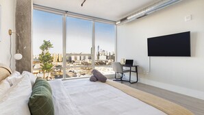 A modern hotel room with a large bed, a desk with a laptop, a TV mounted on the wall, and a view of a cityscape through large windows. - Chique Loft 10 Min from NYC with Gym, Pool, Sauna (Jersey City)