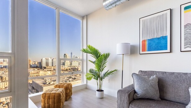 A modern living room with a large window offering a city view, a grey sofa, a floor lamp, and a potted plant. - Chique Loft 10 Min from NYC with Gym, Pool, Sauna (Jersey City)