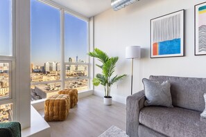 A modern living room with a large window offering a city view, a grey sofa, a floor lamp, and a potted plant. - Chique Loft 10 Min from NYC with Gym, Pool, Sauna (Jersey City)