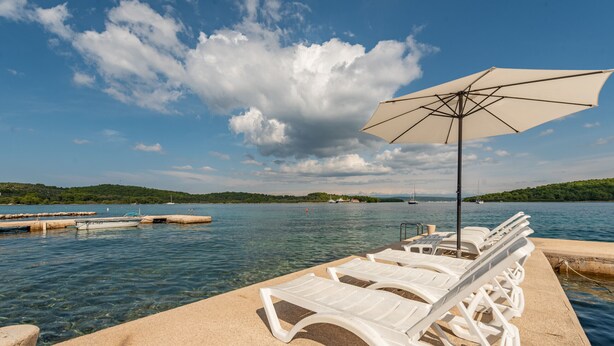 Un espace aménagé au bord de la piscine, avec des chaises longues et un parasol, d’où l’on peut admirer un plan d’eau avec des bateaux, et où la verdure forme un décor en arrière-plan.