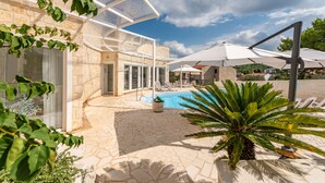 Un espace piscine avec des chaises longues, un patio fermé par des baies vitrées et un bâtiment en pierre.