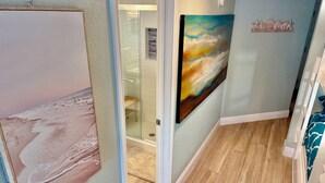 A hallway with a glass door leading to a bathroom, a painting of a beach scene, and a decorative wall piece with shells and starfish. - Seascape 2202 The Seashell Pearl Beachfront Condo Gulf View Pool Hottubs Netflix (Galveston)