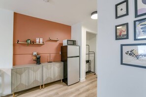 Een kitchenette met een magnetron, koelkast en koffiezetapparaat, geplaatst op een kastje. Aan de muur hangen ingekaderde foto's en er staat een plank met decoratieve objecten.