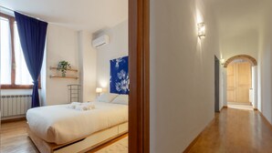 Une chambre avec un lit, une fenêtre avec des rideaux, un radiateur, un climatiseur fixé au mur, un luminaire fixé au mur, une plante et un couloir menant à une autre pièce.
