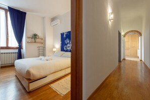 A bedroom with a bed, a window with curtains, a radiator, a wall-mounted air conditioner, a wall-mounted light fixture, a plant, and a hallway leading to another room.