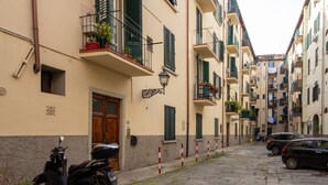 Une rue étroite bordée de voitures garées et d’un scooter, des immeubles dotés de balcons et de volets, et un pavé.