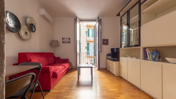 Un espace de vie compact comprenant un canapé rouge, une table à manger avec des chaises, une petite télévision et un coin cuisine avec des placards et une fenêtre.