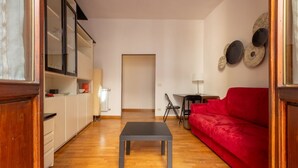 Un espace de vie compact doté d’un plancher en bois, d’un canapé rouge, d’une petite table, d’un coin cuisine avec des placards et d’une porte donnant sur une autre pièce.