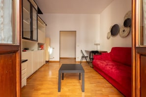A compact living space with a wooden floor, a red sofa, a small table, a kitchen area with cabinets, and a door leading to another room.