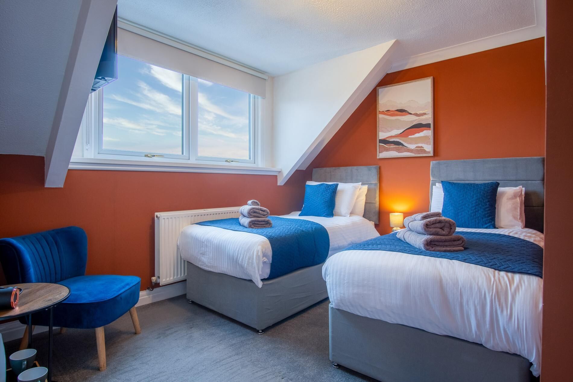 Une chambre d’hôtel moderne avec deux lits, une chaise bleue et une grande fenêtre.